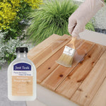 Person applying wood wax oil to a wooden surface with a bottle of Just Teak wood wax oil and a brush.